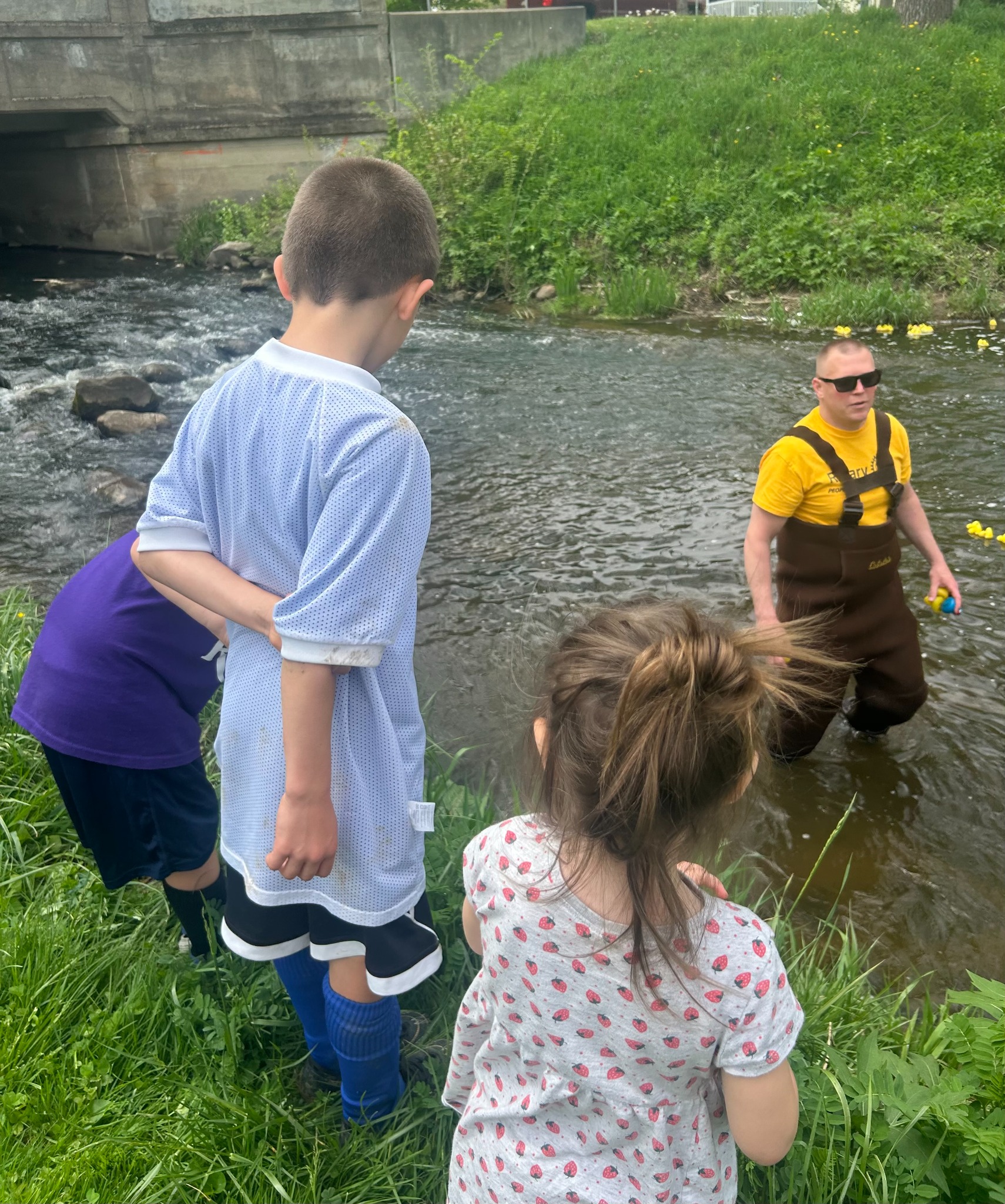 Children watch a community activity beside a creek while an adult stands in the water